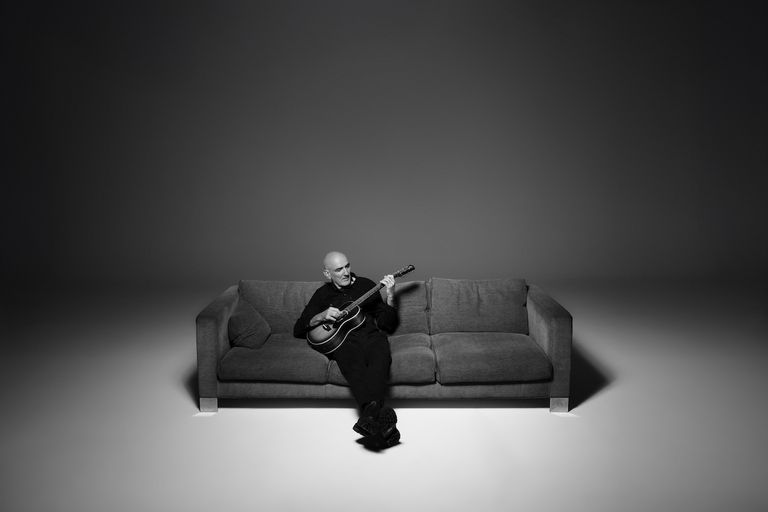 Australian musician Paul Kelly playing acoustic guitar while sitting on a couch in a minimalist black-and-white portrait. Photo by Dean Podmore.